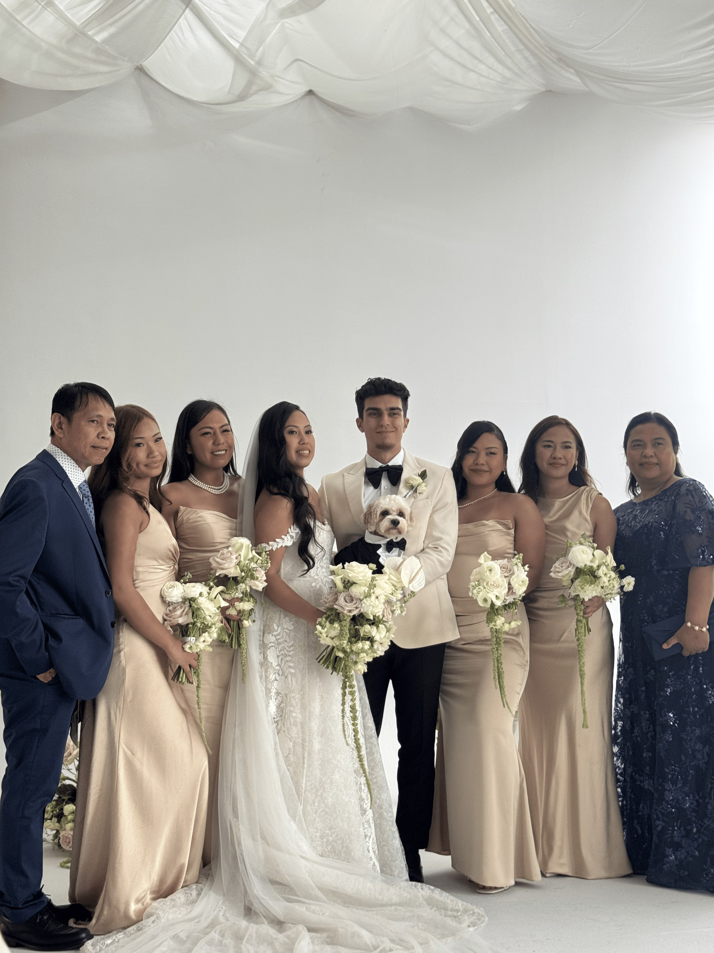 A wedding party in champagne gowns and cream tuxedos against a white draped backdrop, the groom holding a small cream cavapoo in a black bow tie.