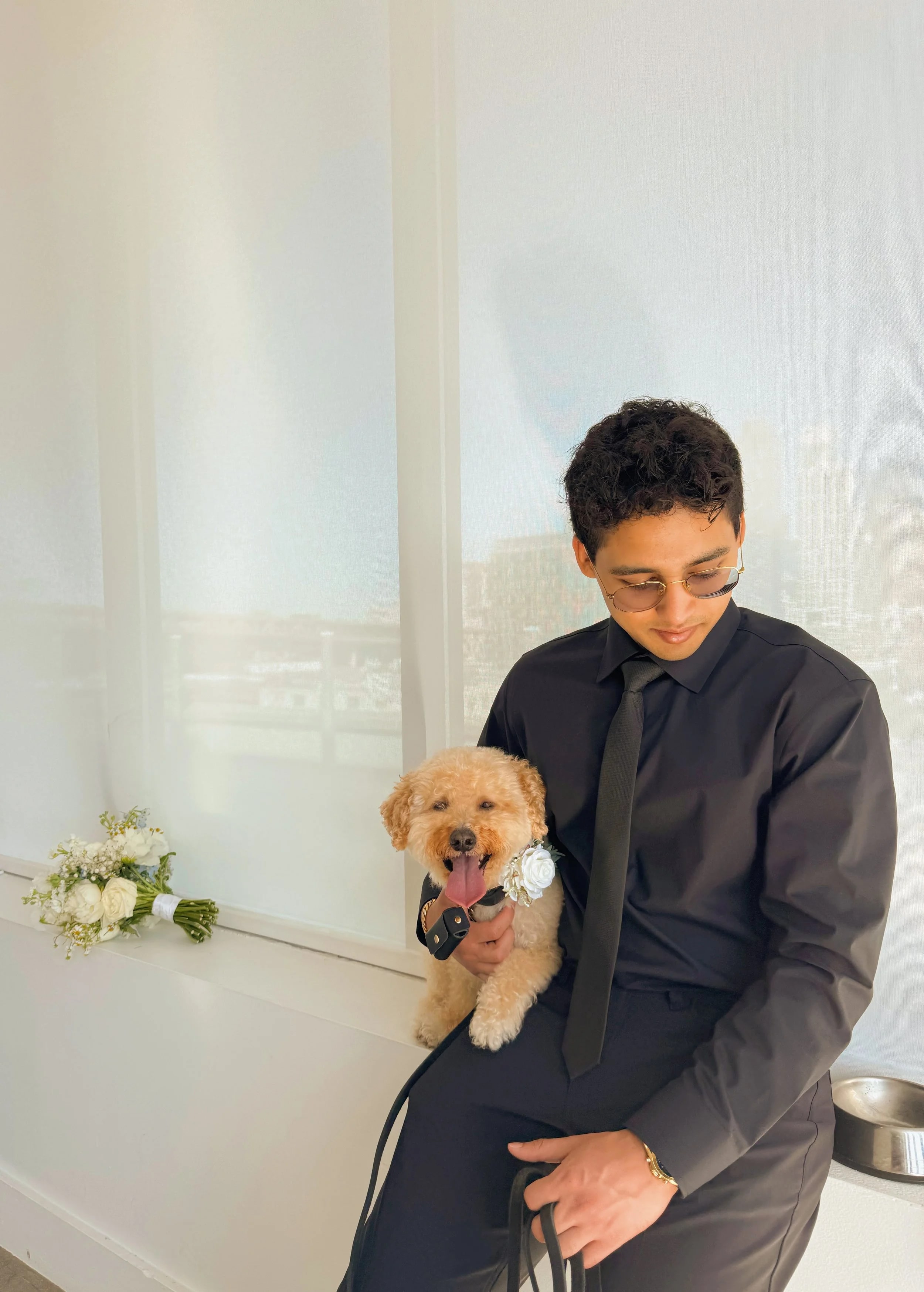An attendant in a black shirt and tie sitting by a window with a tan cavapoo in a white flower collar on his lap, the bridal bouquet resting on the sill.