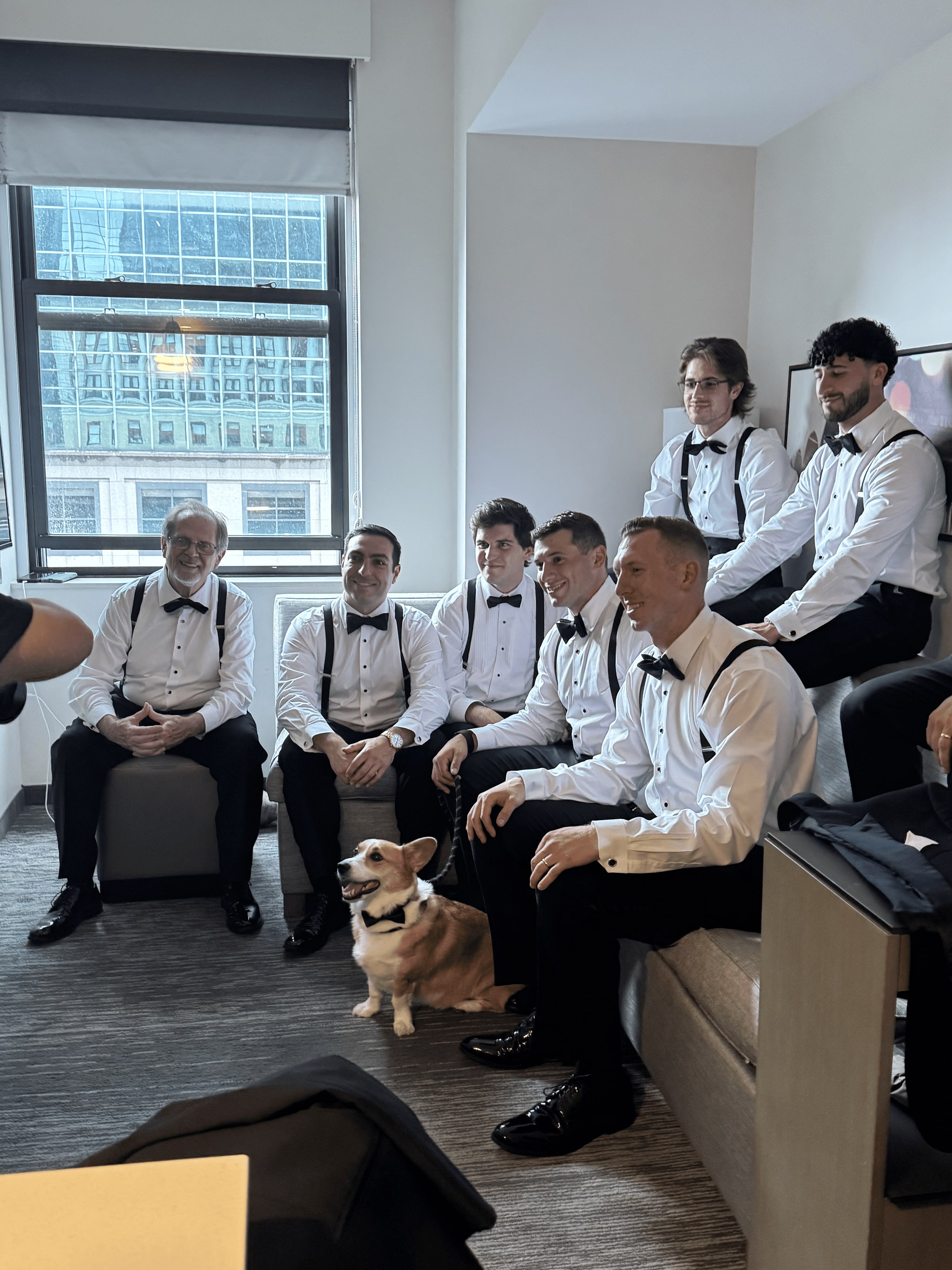 Groomsmen in shirtsleeves, braces and bow ties sitting together in a hotel suite, a corgi in a bow tie waiting on his lead among them.