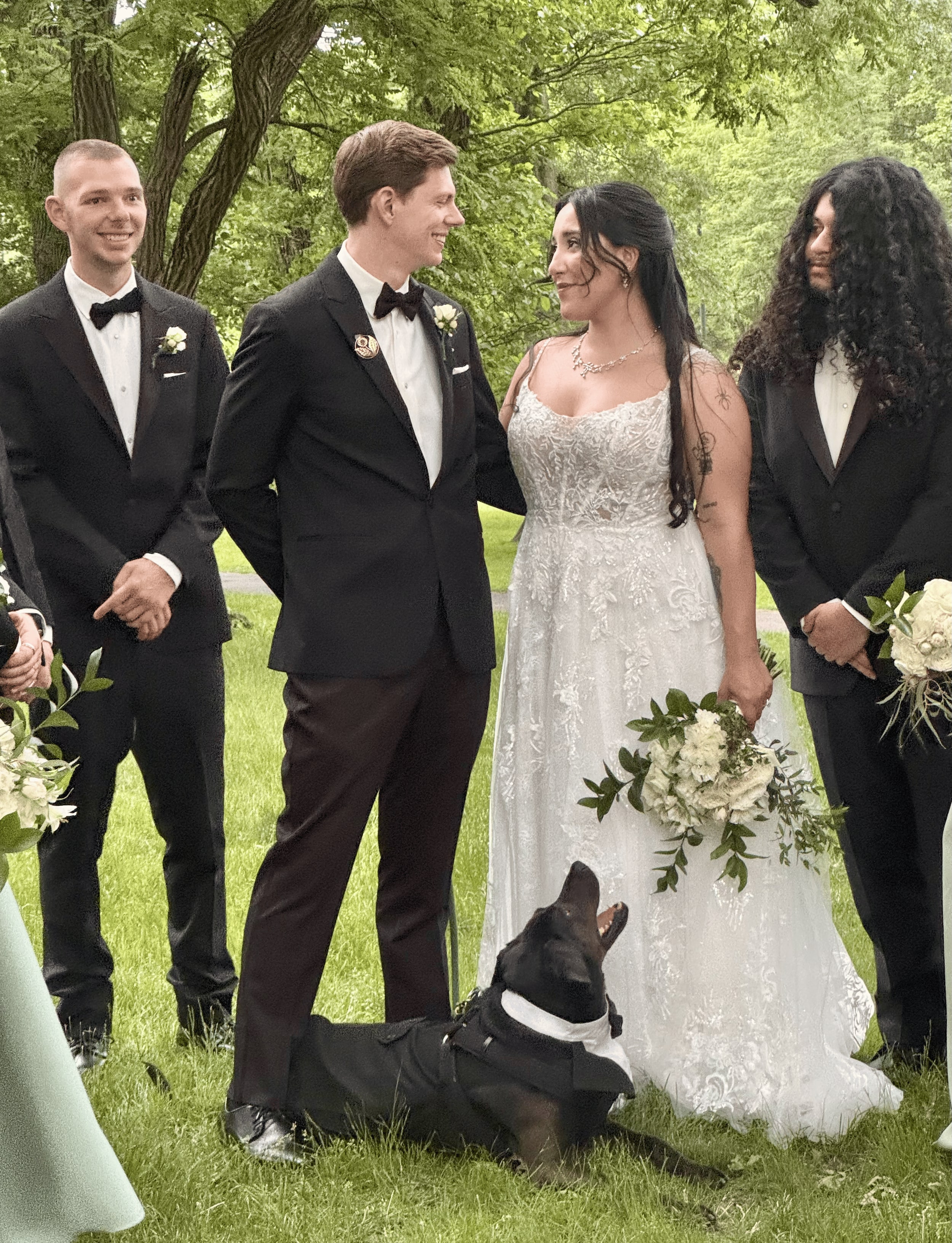 A couple facing each other on a lawn between their groomsmen, a black dog in a tuxedo harness lying at their feet and gazing up at the bride.