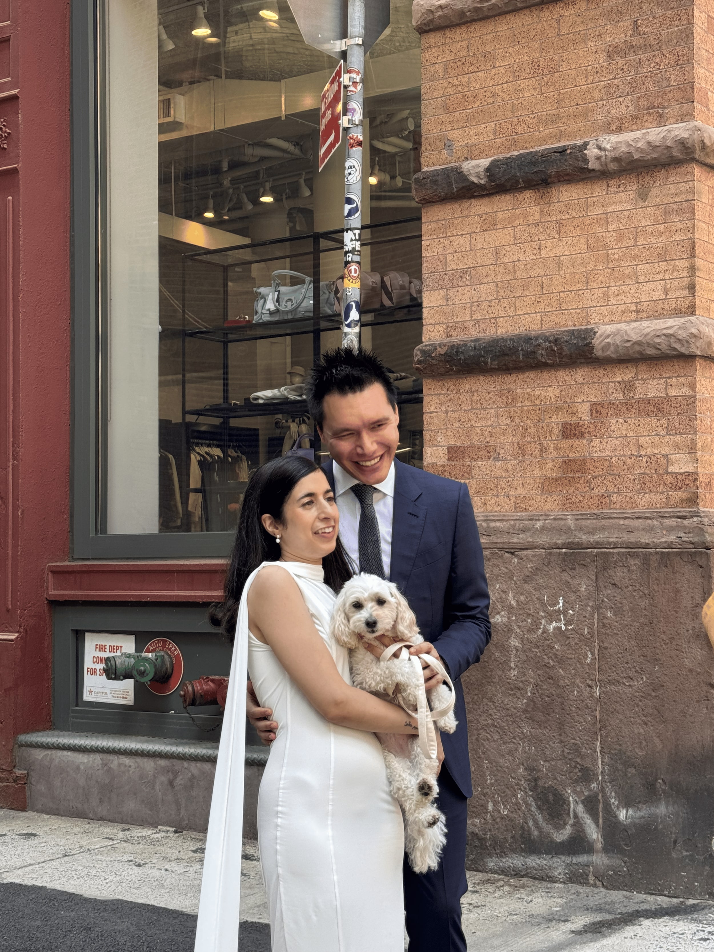 A couple outside a SoHo storefront, the bride in a white gown and the groom in a navy suit cradling a small white curly-coated dog.